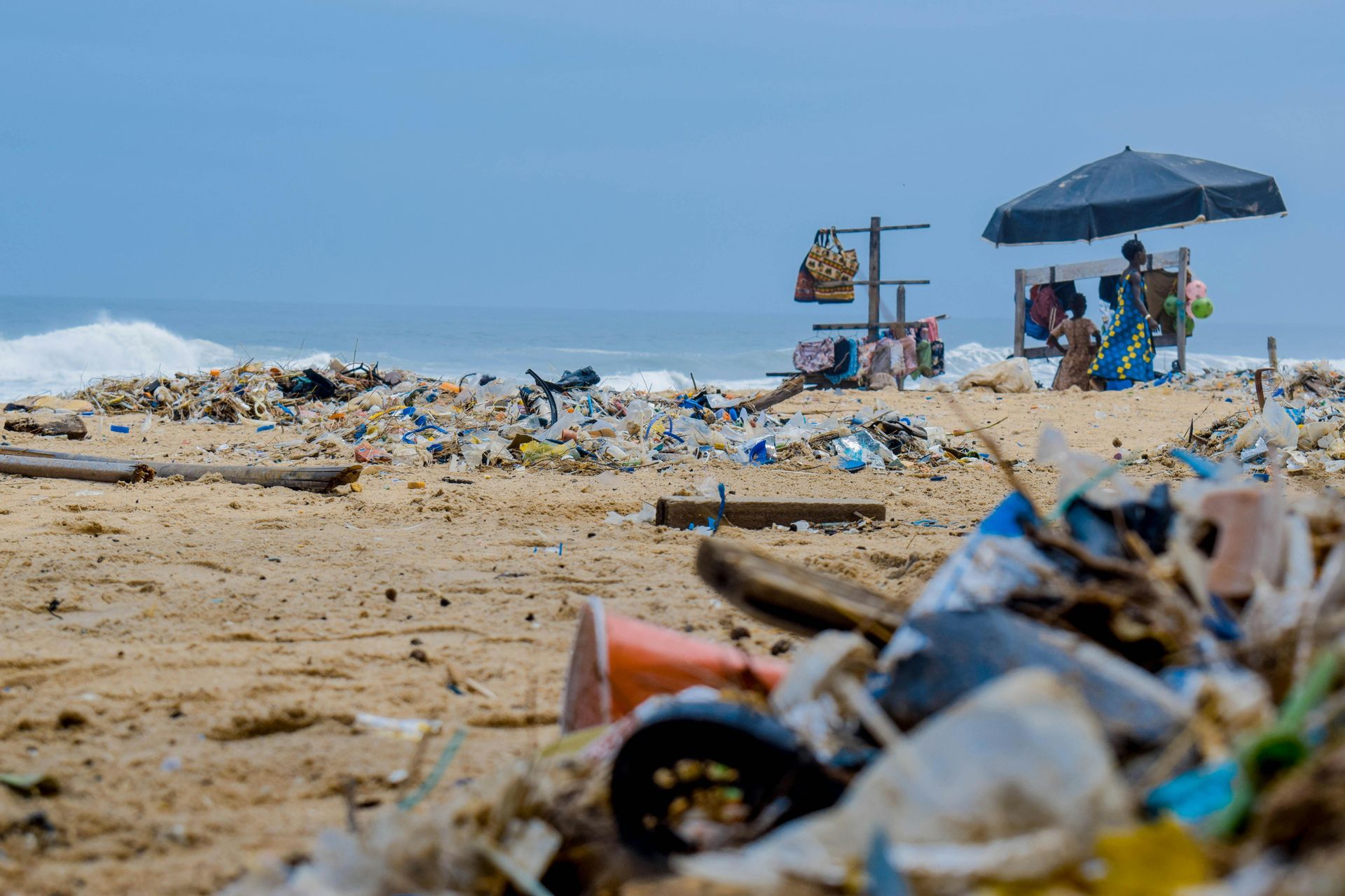 Beach covered in trash; vendor under umbrella sells goods; ocean in background.