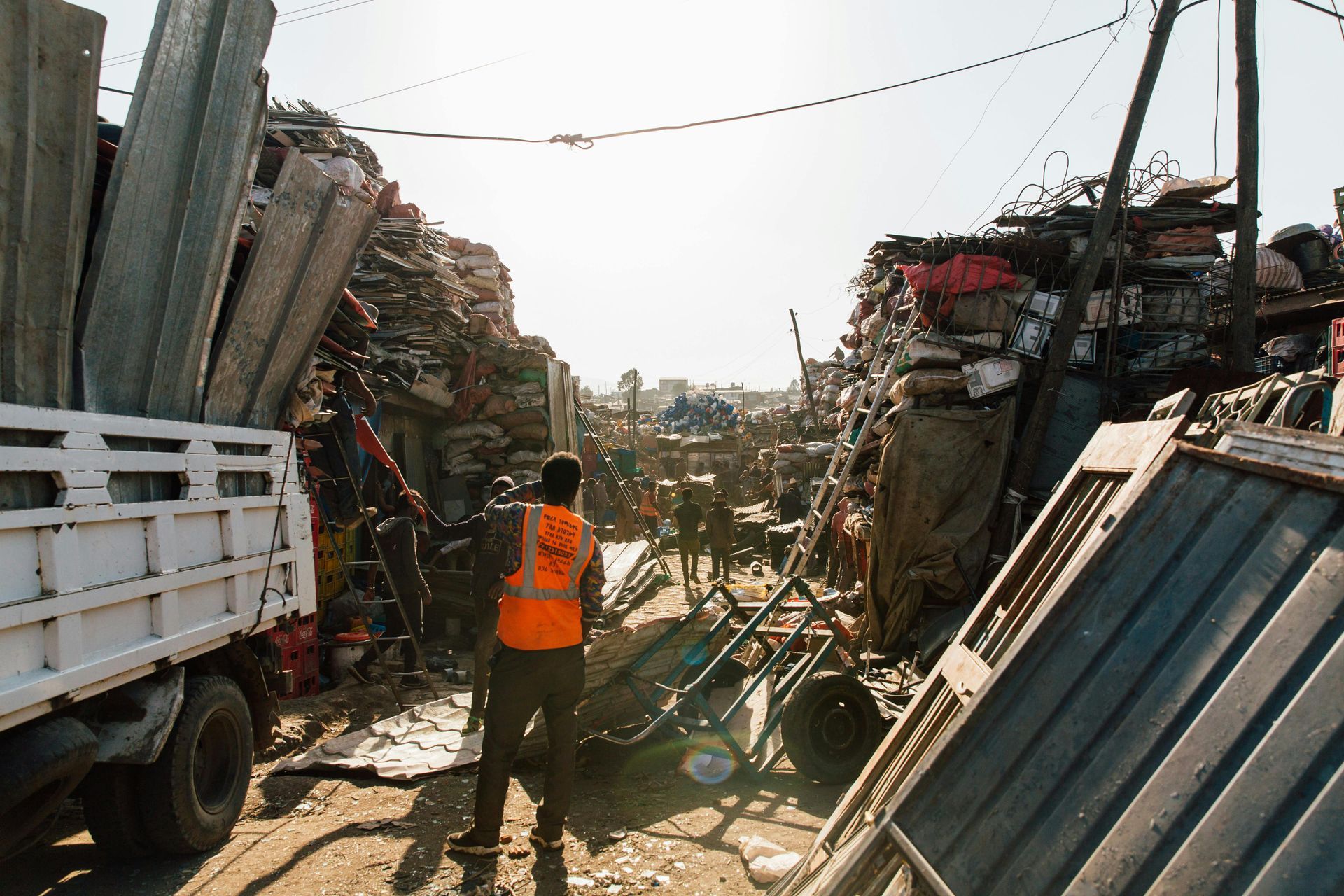 Person in orange vest stands in a cluttered salvage yard with a truck; piles of debris and metal scraps.