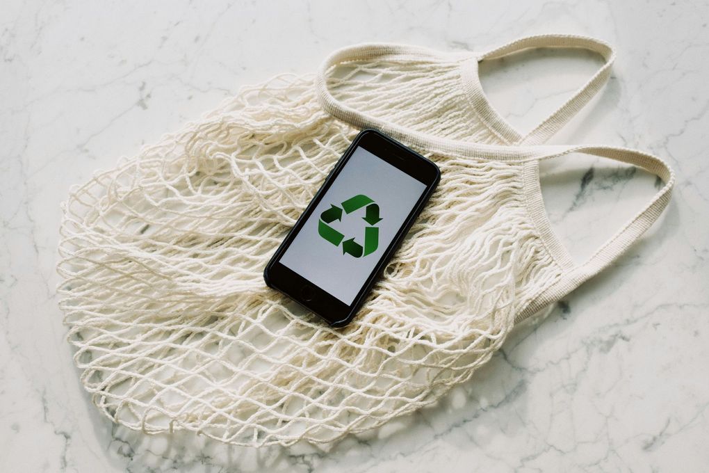Smartphone with recycle symbol on screen rests inside a reusable mesh bag on a marble surface.