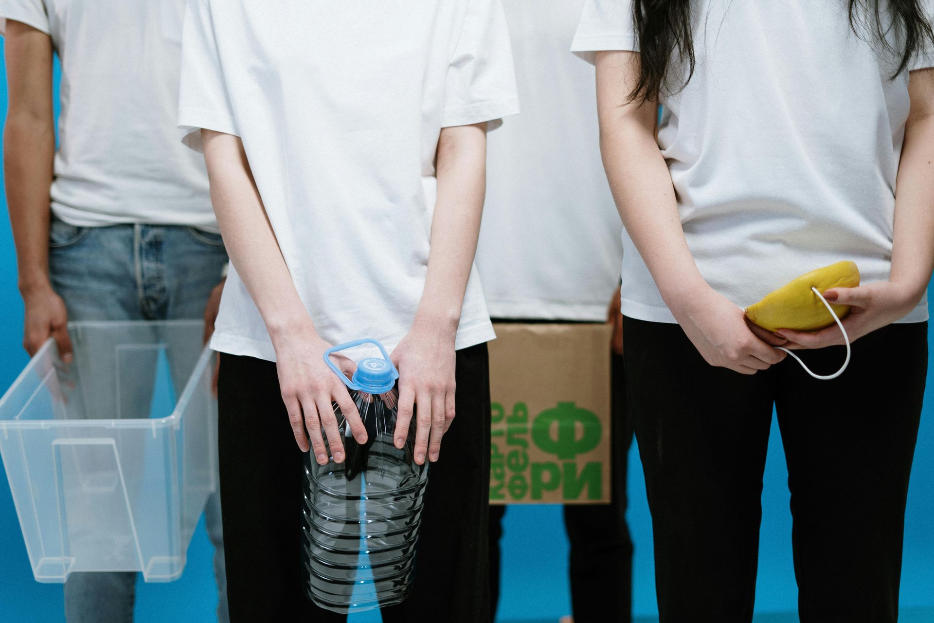 People in white shirts holding recycling bin, bottle, and mask in front of blue background.