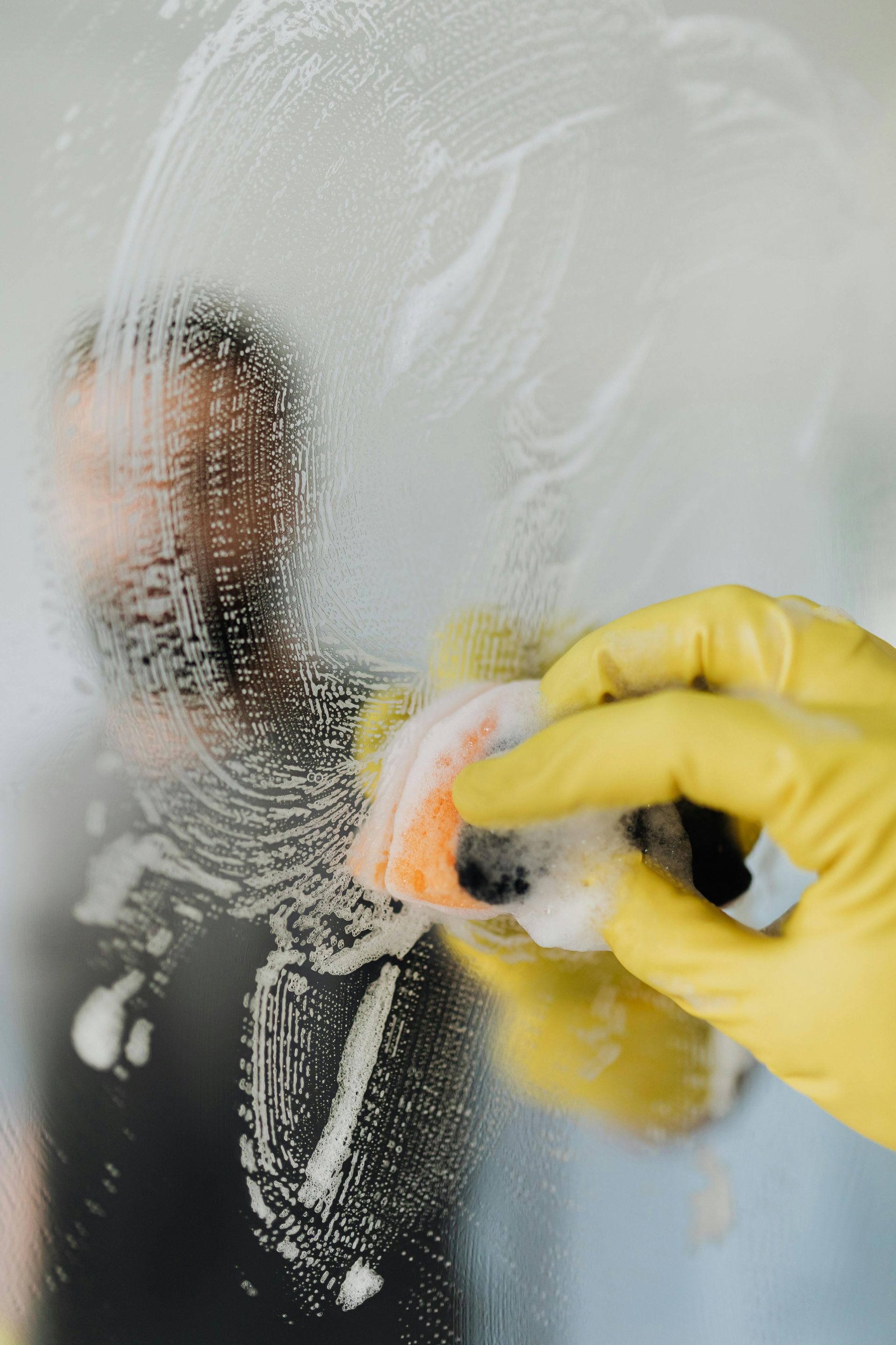A gloved hand scrubs a soapy window, blurring the person's reflection.