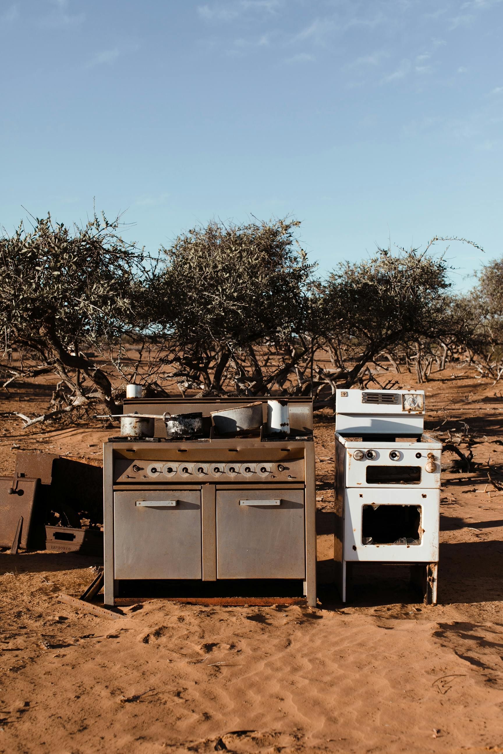 Old kitchen appliances abandoned in a desert, trees and blue sky in the background.