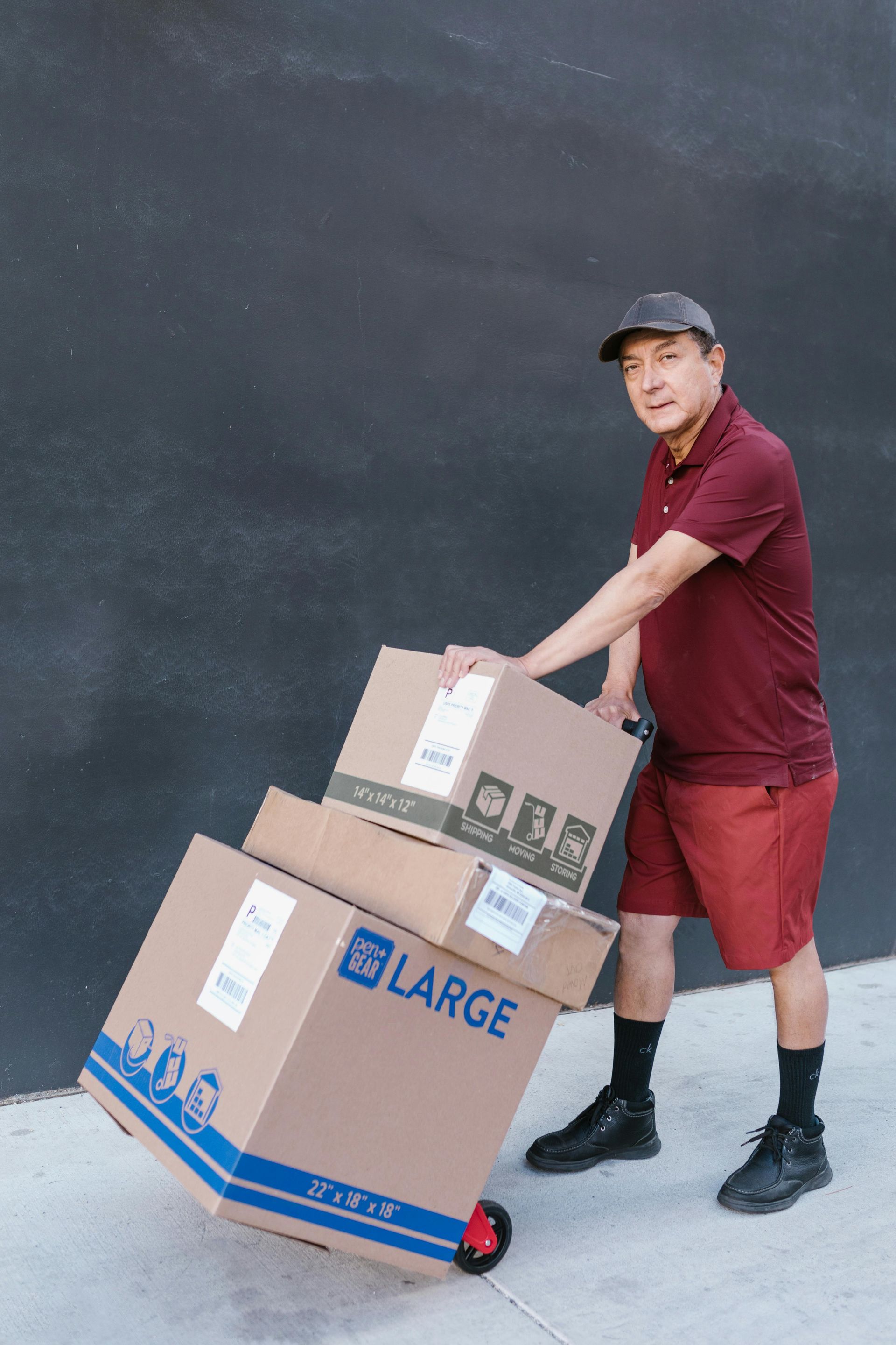 Man in maroon shirt and shorts pulling boxes on a dolly against a dark wall.