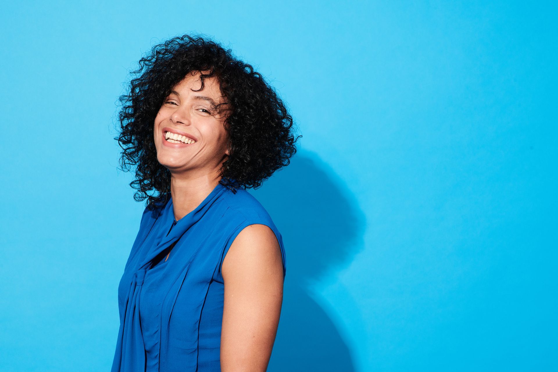 A woman with curly hair is wearing a blue shirt and smiling against a blue background.