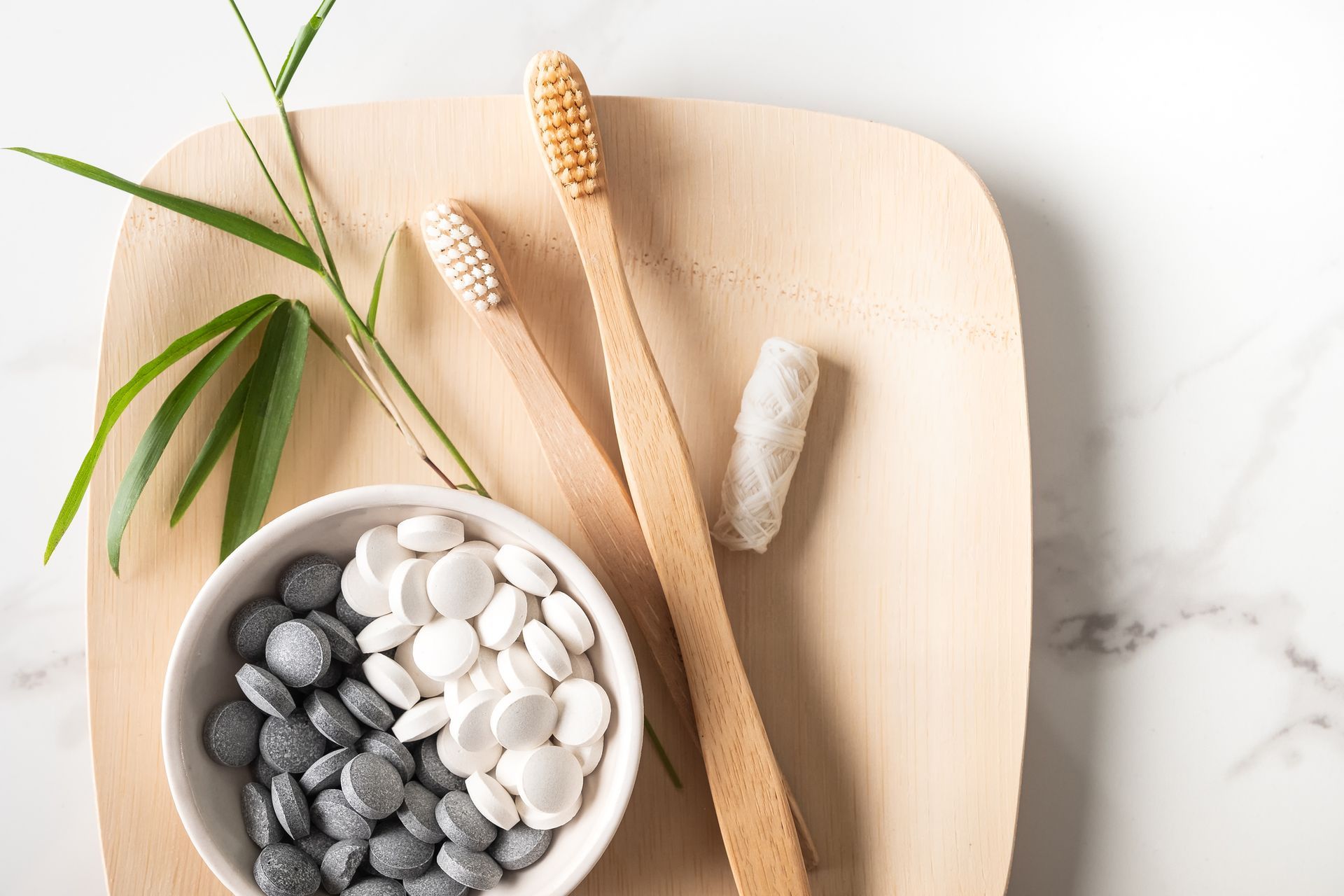 Two wooden toothbrushes and a bowl of toothpaste on a wooden plate.