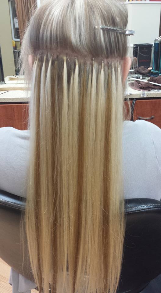 A woman with long blonde hair is sitting in a chair with extensions in her hair.