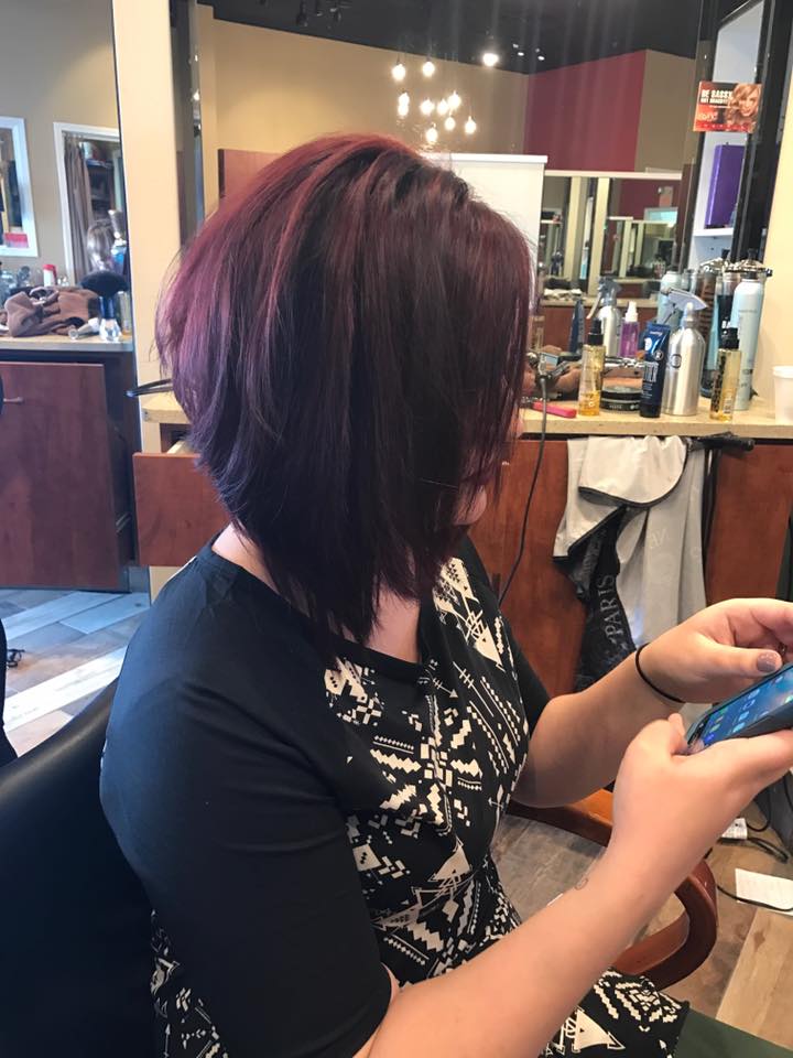 A woman with purple hair is sitting in a chair in a salon holding a tablet.