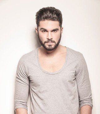 A man with a beard wearing a grey shirt is standing in front of a white wall.