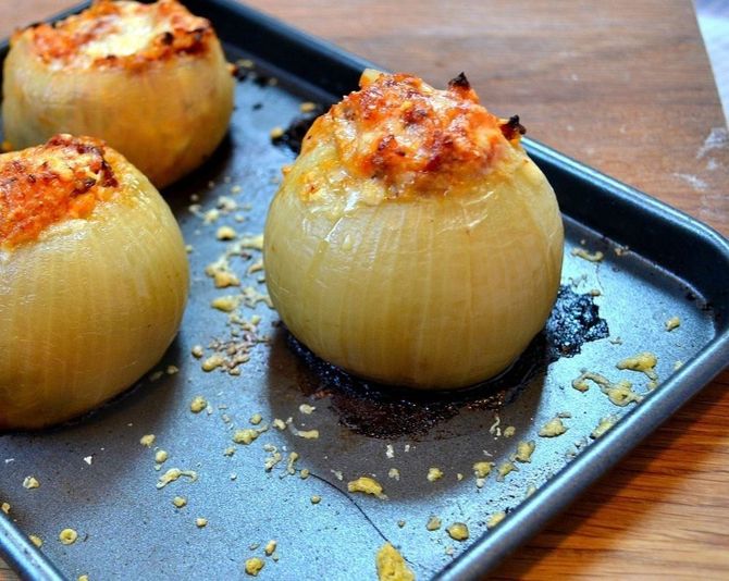 Tre cipolle ripiene cotte al forno su una teglia scura, ricoperte da briciole di formaggio rosolato.