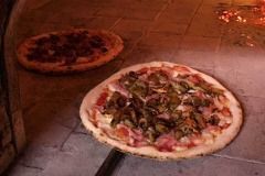 Due pizze cuociono sul fondo di un forno a legna, una davanti con verdure e una semplice dietro.