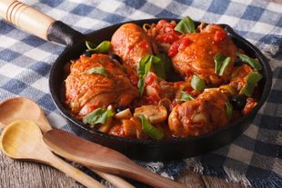 Padella in ghisa con pollo arrosto in salsa e verdure su una tovaglia a quadri blu, con cucchiai di legno nelle vicinanze.