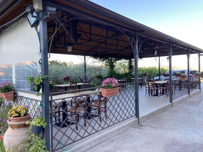 Zona pranzo esterna con patio, arredata con tavoli e sedie in metallo e vasi di fiori rosa, il tutto sotto una tettoia.