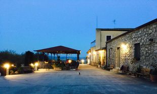 Cortile al crepuscolo con edificio in pietra, vialetto illuminato e patio coperto.