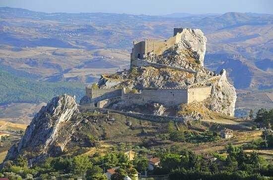 Rovine di un castello in pietra in cima a una collina rocciosa, con vista su un'ampia valle rurale e sulle montagne in lontananza.