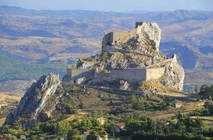 Castello in pietra in rovina su una collina rocciosa che domina un paesaggio arido e montuoso.