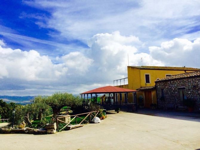 Un soleggiato cortile rurale accanto a un edificio giallo sotto un cielo azzurro con nuvole.