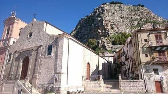 Chiesa in pietra e edifici adiacenti ai piedi di una collina rocciosa, sotto un cielo limpido.