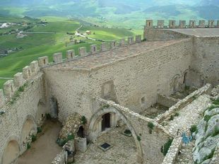 Mura di pietra del castello che si affacciano su una verde vallata, con merlature e un ingresso ad arco sottostante.
