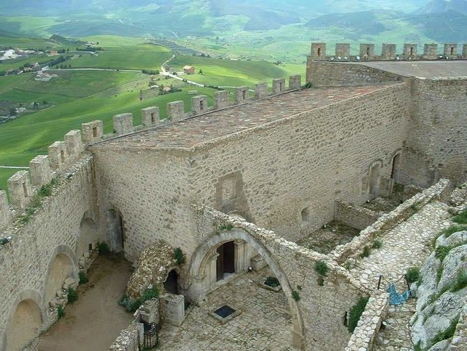 Mura di pietra del castello che si affacciano su una verde vallata, con merlature e un ingresso ad arco sottostante.