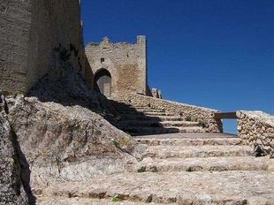 Scalini di pietra conducono a un cancello di castello consumato dal tempo, su un pendio roccioso sotto un cielo azzurro e limpido.