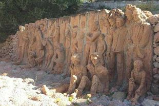 Grande scultura in argilla raffigurante figure umane allineate contro un muro all'aperto, in tonalità marroni terrose.