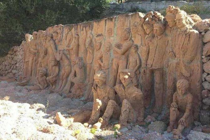 Grande scultura in argilla raffigurante figure umane allineate contro un muro all'aperto, in tonalità marroni terrose.