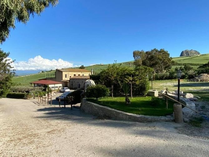Patio di una casa colonica rurale con ombrelloni rossi, auto parcheggiata e verdi colline sotto un cielo azzurro.