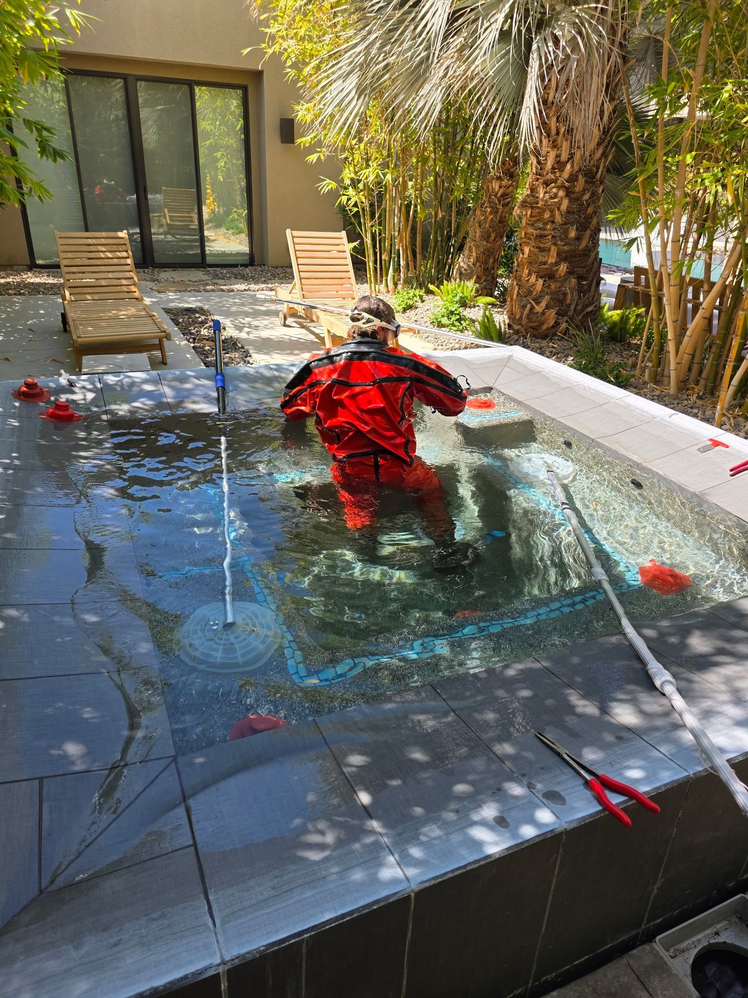 Person in a red shirt sits in a shallow rooftop pool beside lounge chairs and a palm tree.