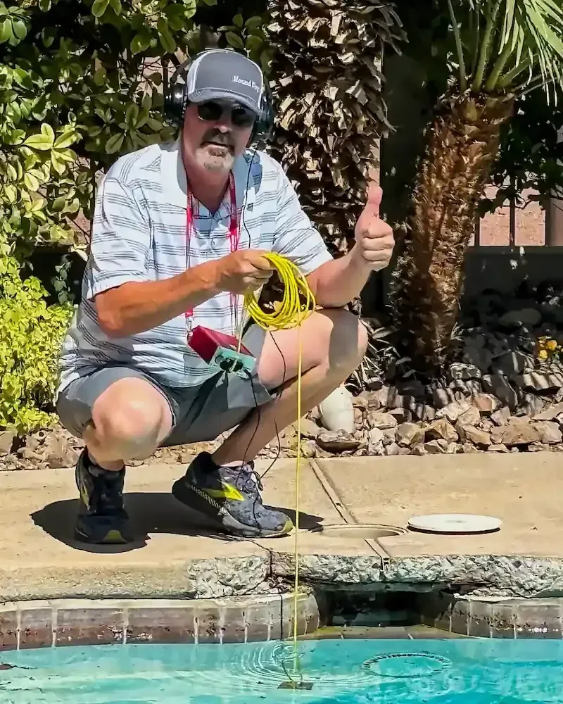 Man kneeling by a pool, holding a yellow rope and giving a thumbs-up near a yellow cord in the water