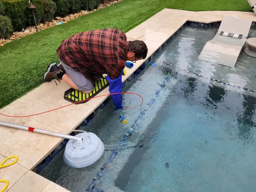 Person kneeling at the edge of a pool, cleaning the water with a hose and equipment