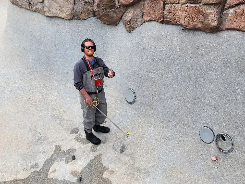 Person standing in a drained pool holding a tool, with pool drains and a rocky edge nearby