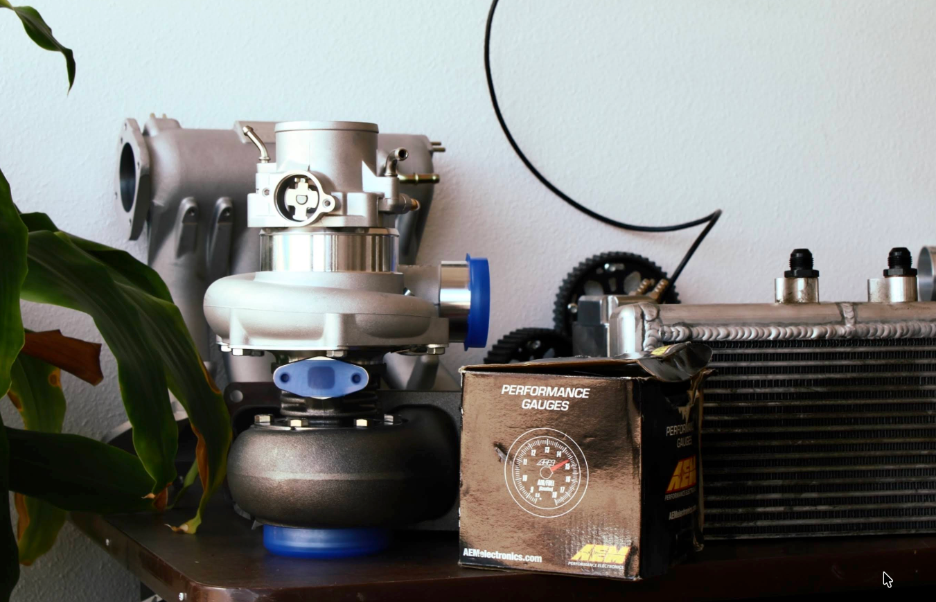 A turbocharger, intercooler, and box sit on a surface next to a plant, against a white wall. | Jeanithe Motorsports