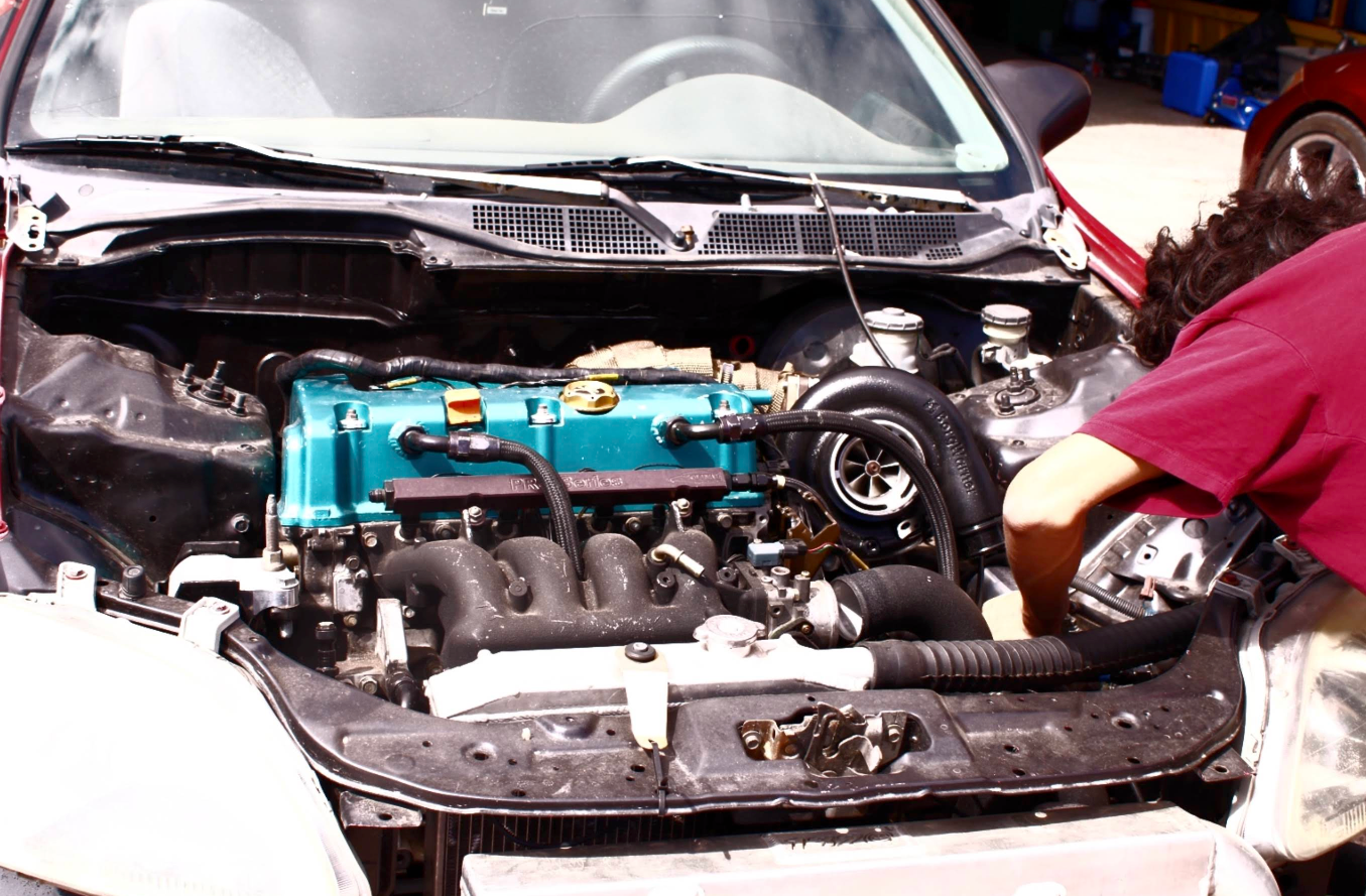 Person working on the engine of a car. Blue engine cover, black tubing, person wearing red shirt. | Jeanithe Motorsports