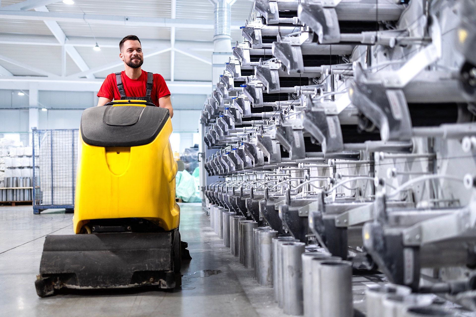A worker in a red shirt operates a bright yellow industrial floor scrubber in a warehouse with metal machinery shelving.
