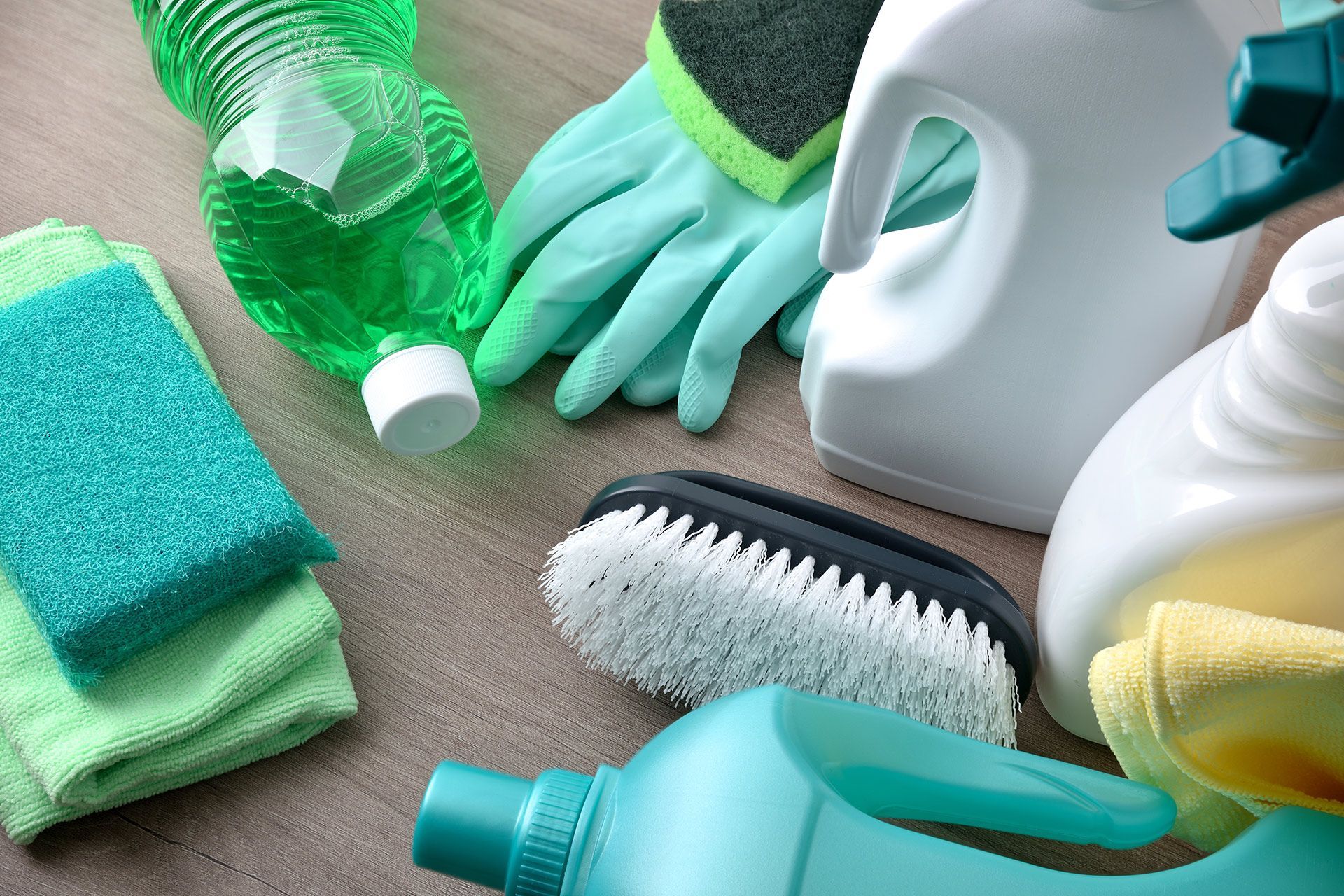 A variety of cleaning supplies, including gloves, sponges, brushes, and bottles of cleaner, arranged on a wooden surface.