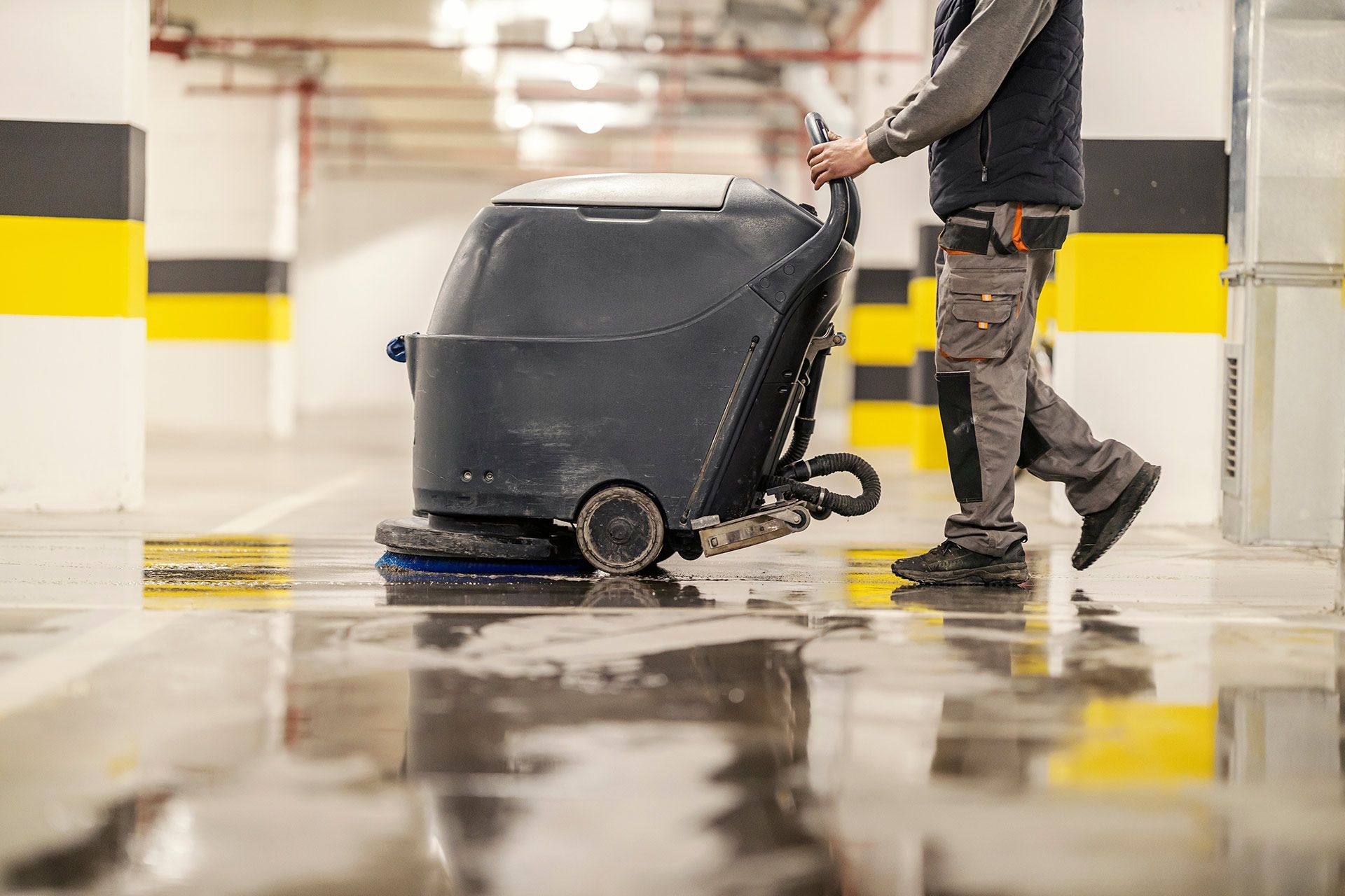 A worker uses a walk-behind floor scrubber to clean a wet parking garage with yellow and black painted pillars.