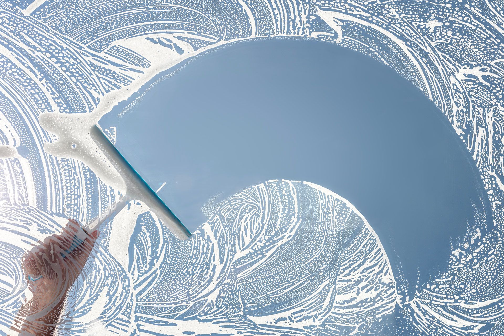 A squeegee held by a hand clears a streak of soapy foam off a glass window, revealing a clear blue sky behind.