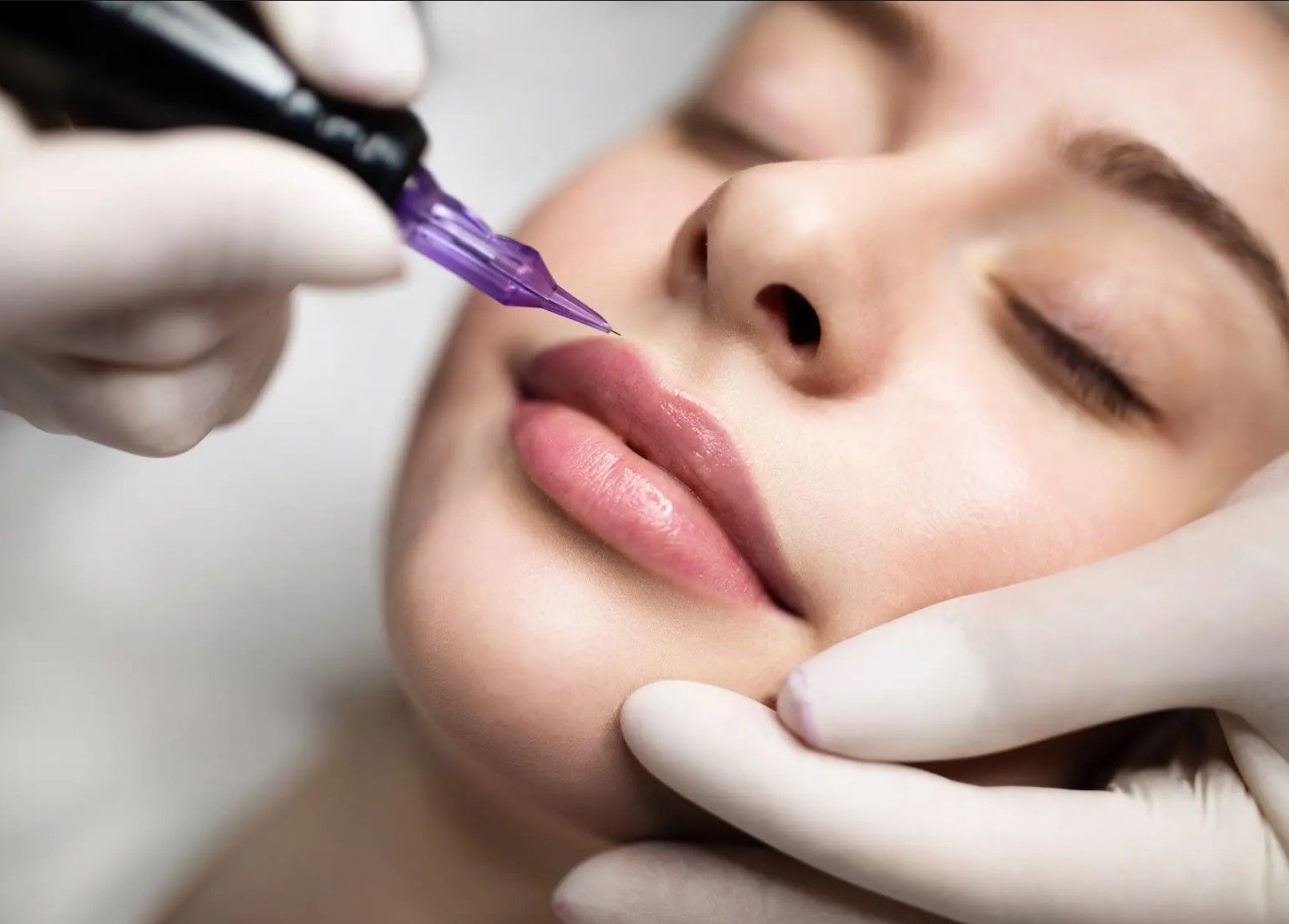 A woman receiving lip tattoo, pale pink lips, purple needle, white gloves, and closed eyes.