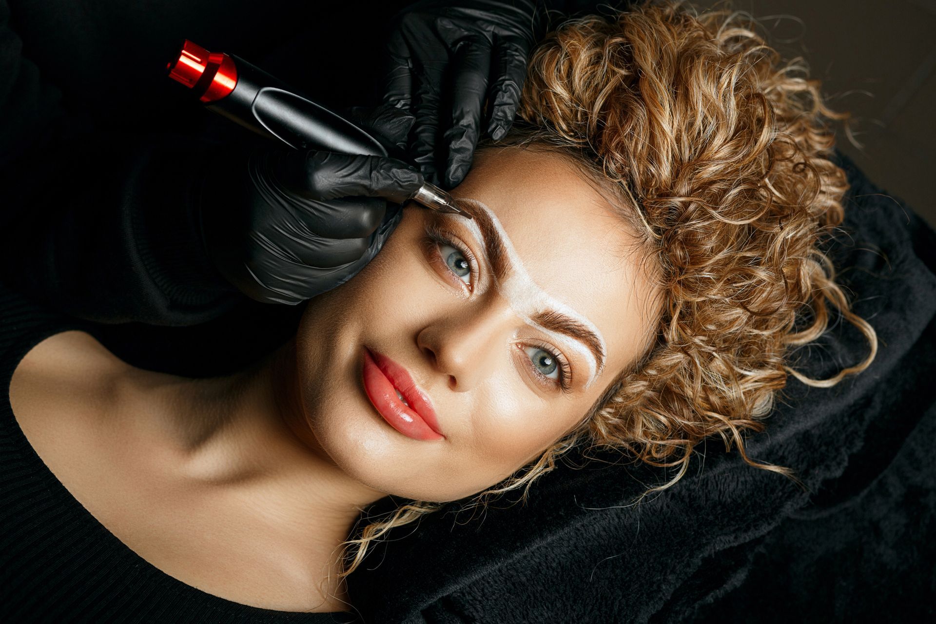 Woman getting eyebrow microblading. A technician in black gloves uses a tool to create a brow shape.