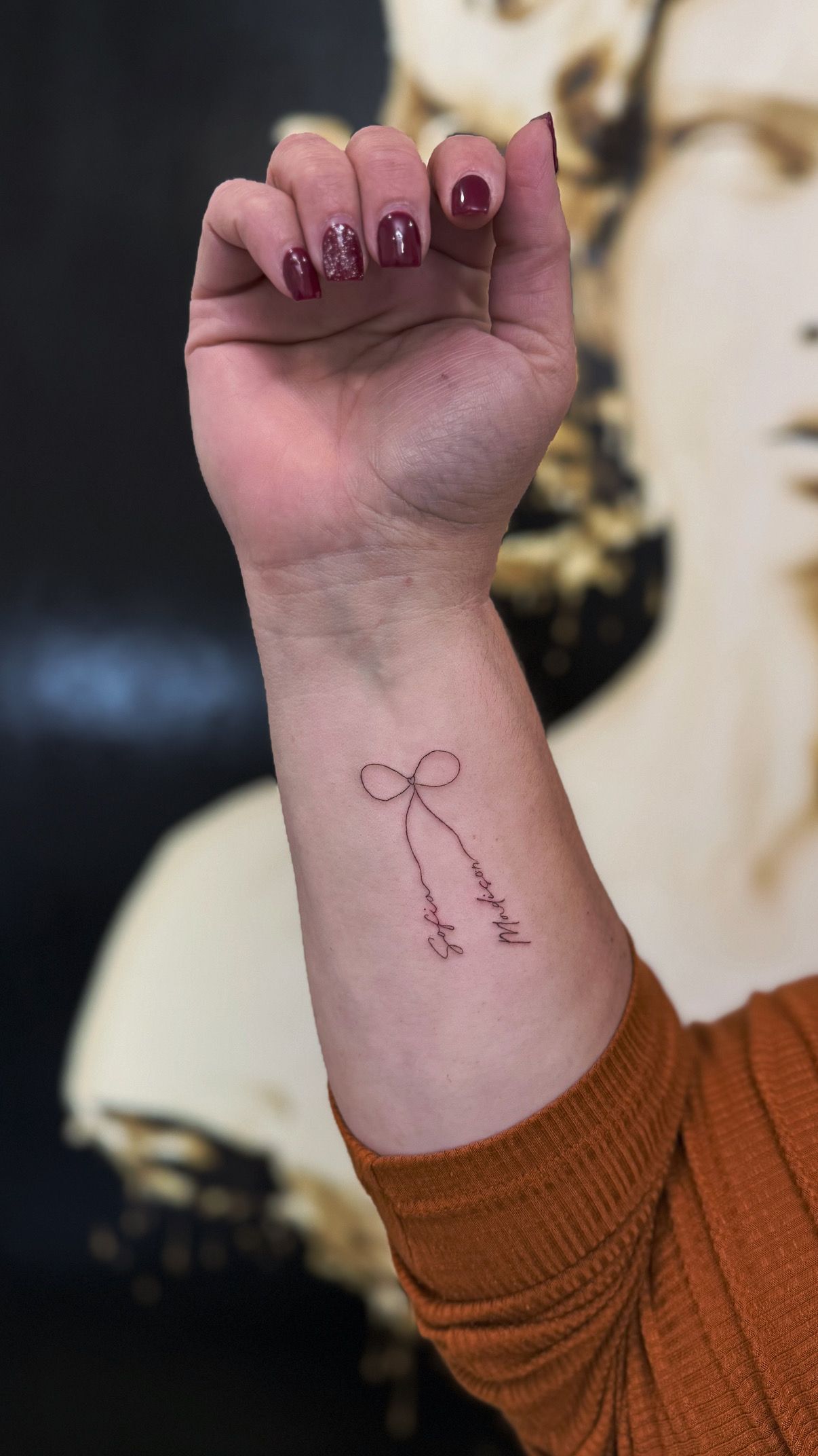 A person's forearm showing a small, fine-line tattoo of a bow with hanging ribbons, set against a blurred background.