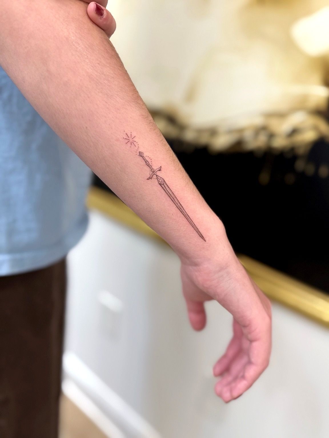 A close-up of a forearm featuring a fine-line tattoo of a slender sword with a small sparkling star above its hilt.