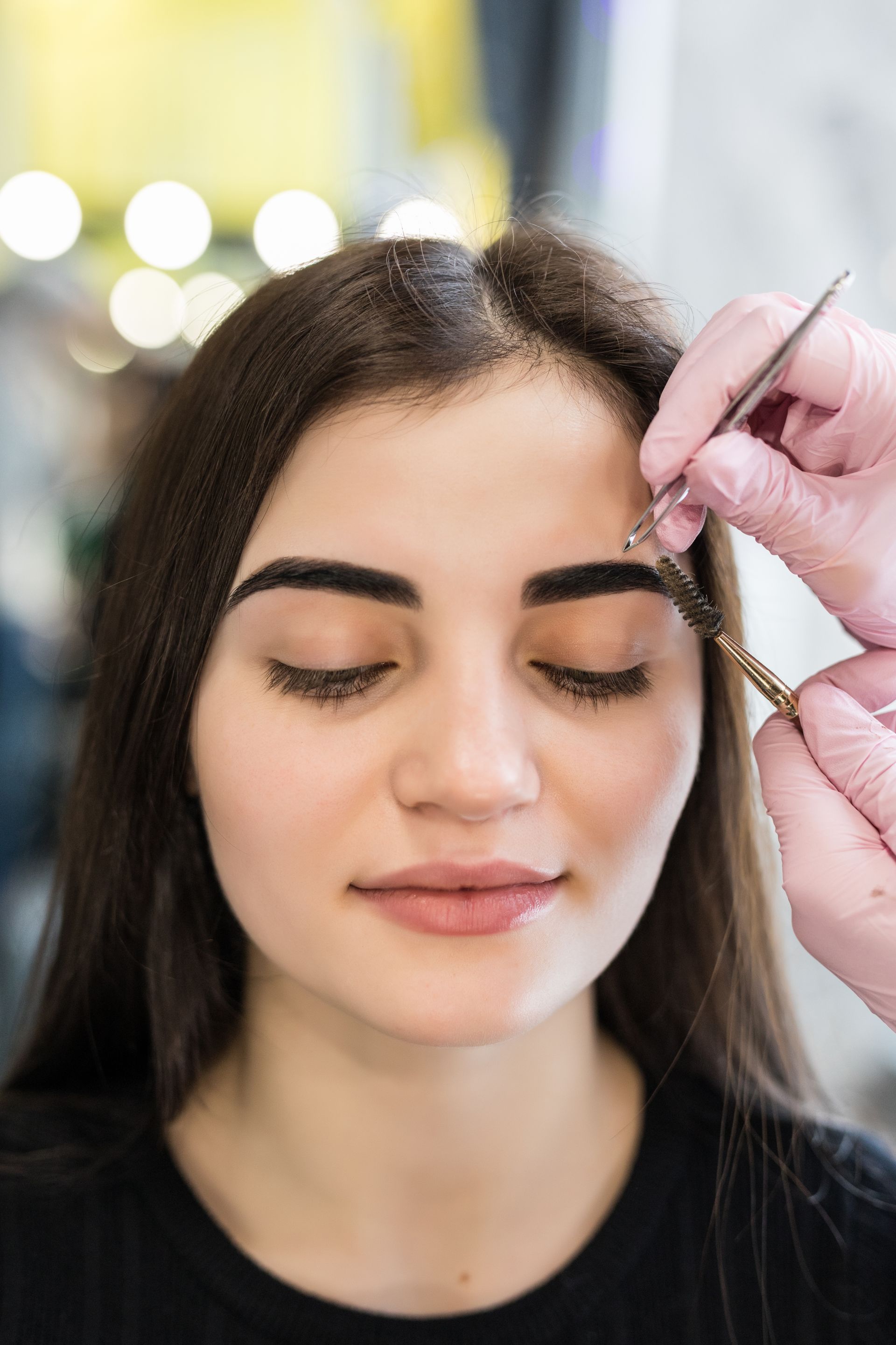 Woman with closed eyes has her eyebrow being brushed by someone with gloved hands.