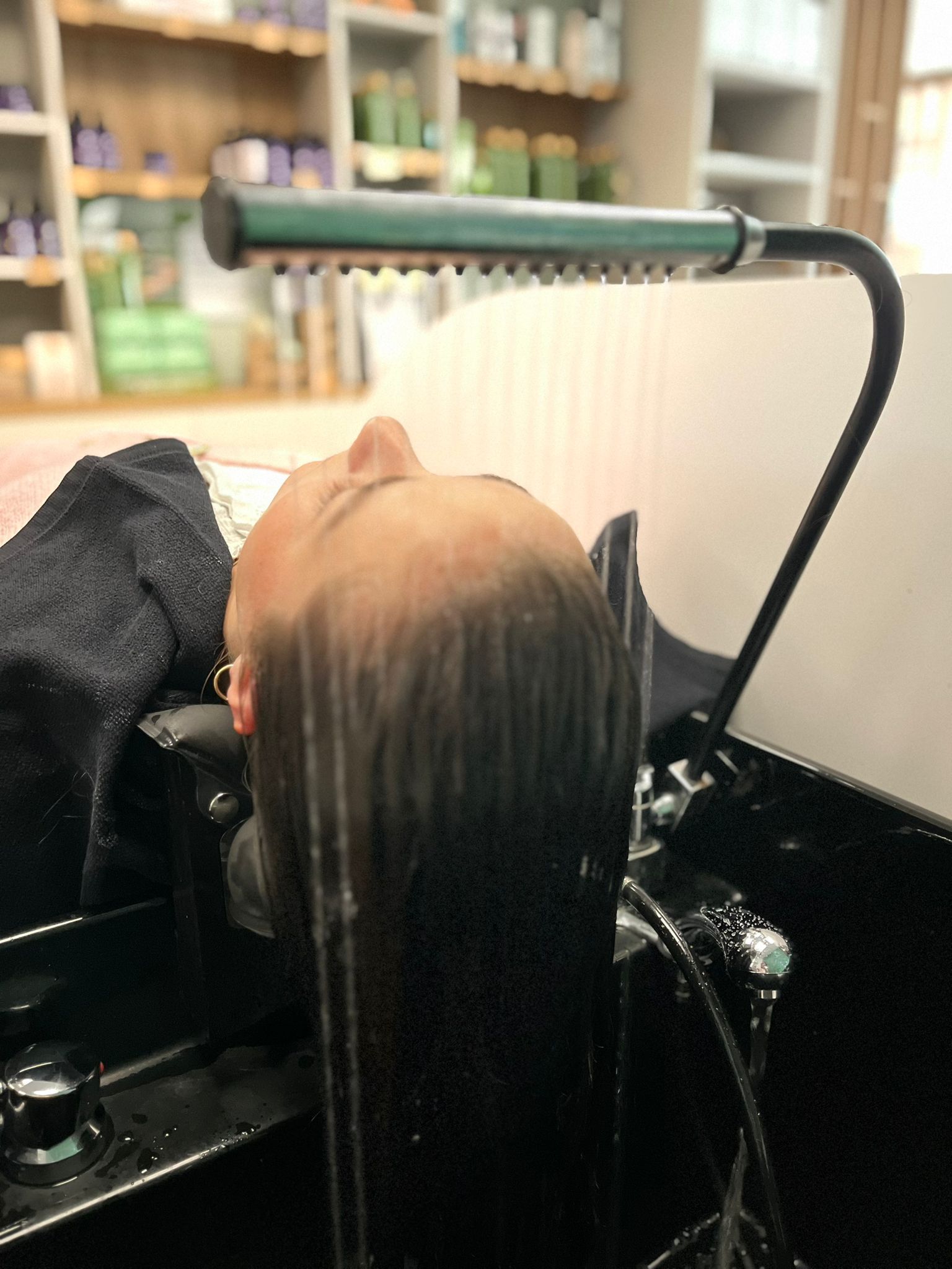 Une personne se fait laver les cheveux dans un salon. L'eau coule sur ses cheveux depuis un pommeau de douche vert.