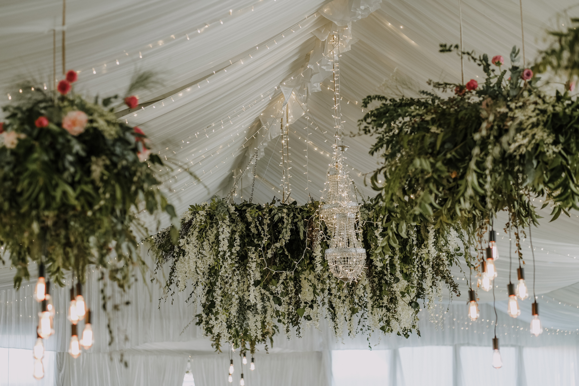 Wedding tent interior with hanging floral arrangements, fairy lights, and exposed light bulbs.