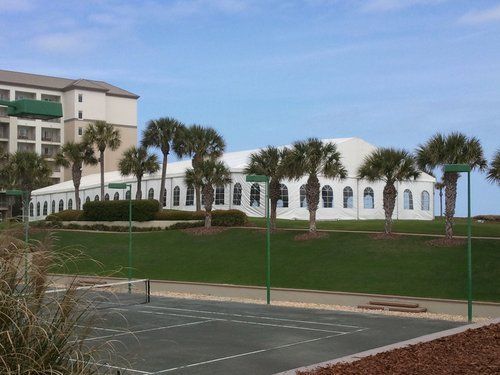 Tennis court in foreground with a white building and palm trees on a grassy hill under a blue sky. A tall building is on the left.