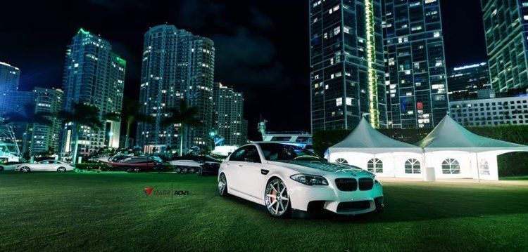 White BMW sedan parked on a lawn at night, with tall buildings illuminated in the background.