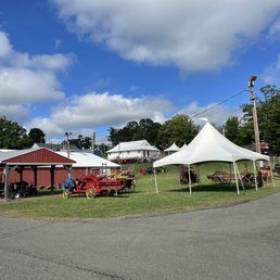 A fairground with white tents and red agricultural equipment under a blue sky with puffy clouds.