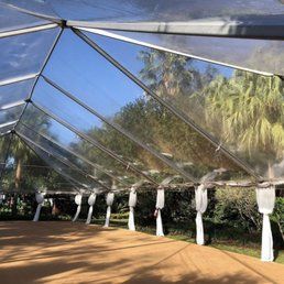 Inside view of a clear tent, revealing the sky and foliage. White fabric drapes line the tent poles along the side.