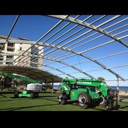 Two green construction vehicles working under a metal frame structure on a grassy field, with a building and water in the background.