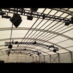 Inside of a large white tent; black speaker systems are mounted on suspended metal beams.